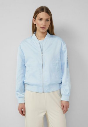 Kobieta w jasnoniebieskiej bomberce zapinanej na zamek i kremowych spodniach, stojąca na tle jednolitego szarego tła.