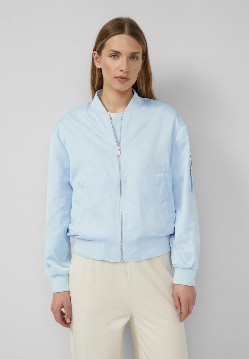 Žena oblečená v svetlo modrej bomber bunde na zips a krémových nohaviciach, stojaca pred jednoduchým sivým pozadím.