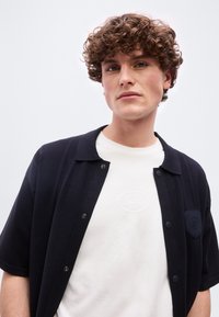 Νεαρός με σγουρά μαλλιά που φοράει λευκό t-shirt με ανάγλυφο λογότυπο κάτω από μαύρο κοντομάνικο πουκάμισο με κουμπιά, μπροστά σε απλό φόντο.