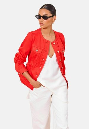 Femme aux cheveux plaqués en arrière portant des lunettes de soleil noires, une veste rouge boutonnée, un haut blanc col en V et un pantalon blanc avec une main dans la poche.