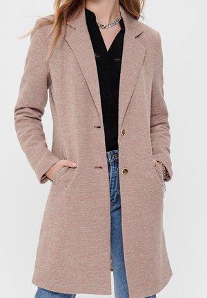 Kobieta w beżowym, teksturowanym, długim płaszczu z guzikami, czarnej bluzce, niebieskich dżinsach i srebrnym łańcuszku na szyję, z rękami w kieszeniach płaszcza.