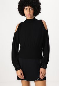 Black knitted sweater featuring cold shoulder cutouts, long sleeves, a high neckline, and textured beaded cuffs. Form-fitting waist.