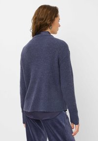 Cardigan a maglia blu navy con texture a coste e apertura frontale. Maniche lunghe e dettagli sulla cucitura posteriore. Indossato sopra un top scuro e abbinato a pantaloni.