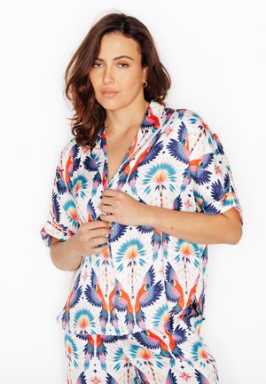 Donna con capelli castani fino alle spalle che indossa una camicia a maniche corte con bottoni e pantaloni coordinati con motivo di pappagalli colorati, mentre sistema la parte anteriore della camicia.