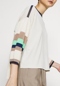 Krämvit tröja med ribbade ärmar i flera färger: brun, mintgrön och mörkblå. Har rund halsringning och ledig passform.