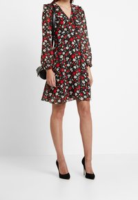 Svart blommig klänning med röda och vita blommor, genomskinliga långärmade ärmar, V-urringning och flödande kjol. Stylad med svarta högklackade skor och en liten väska.