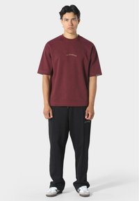 Burgundský krátký rukáv svetr s nápisem "PAS DE MONACO" tištěným zlatou barvou, v kombinaci s volnými černými kalhotami a bílými teniskami.