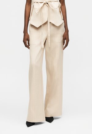 Žena nosi béžové nohavice širokého strihu s prednými vreckami, doplnené čiernymi topánkami s špicatou špičkou, stojaca pred jednoduchým pozadím.