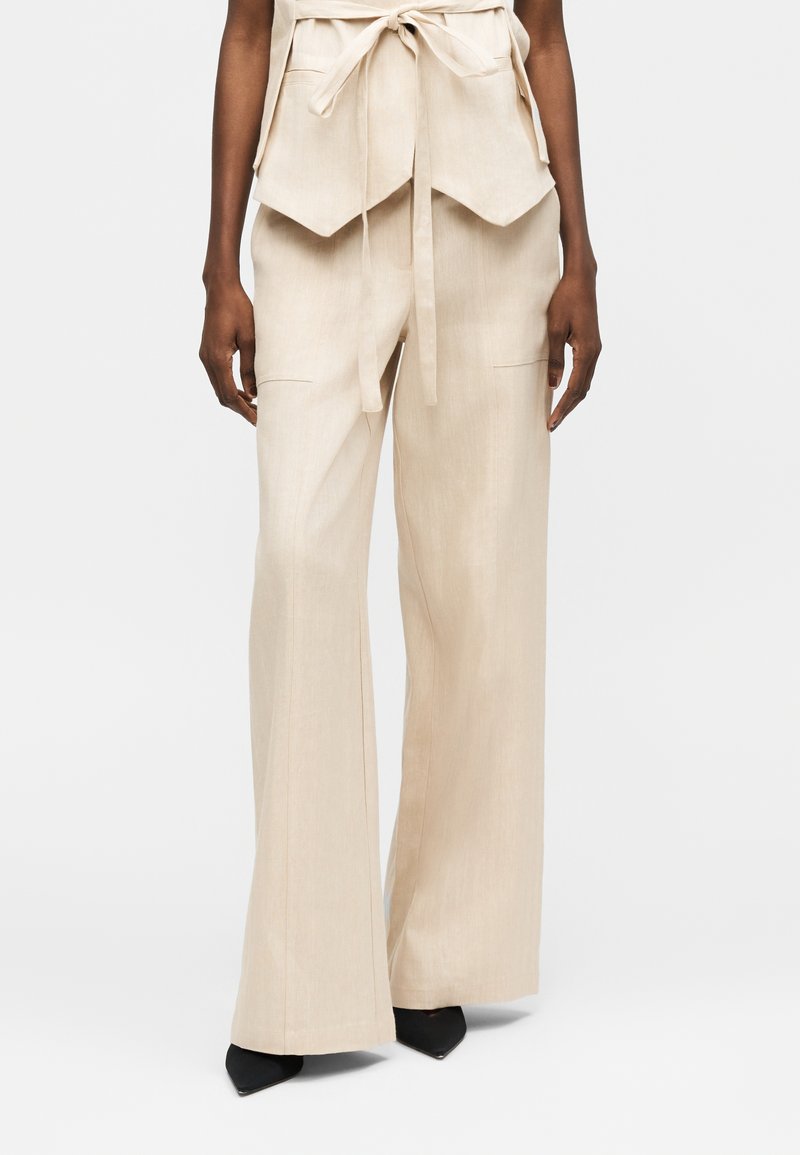 Γυναίκα που φοράει μπεζ παντελόνια με φαρδύ πόδι και μπροστινές τσέπες, σε συνδυασμό με μαύρα παπούτσια με μυτερή μύτη, στέκεται μπροστά σε απλό φόντο.