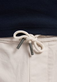 Mørkeblå ribbet stoftop overlapper beige stofbukser, med en hvid snor knyttet i en sløjfe med metalender mærket "PETROL."