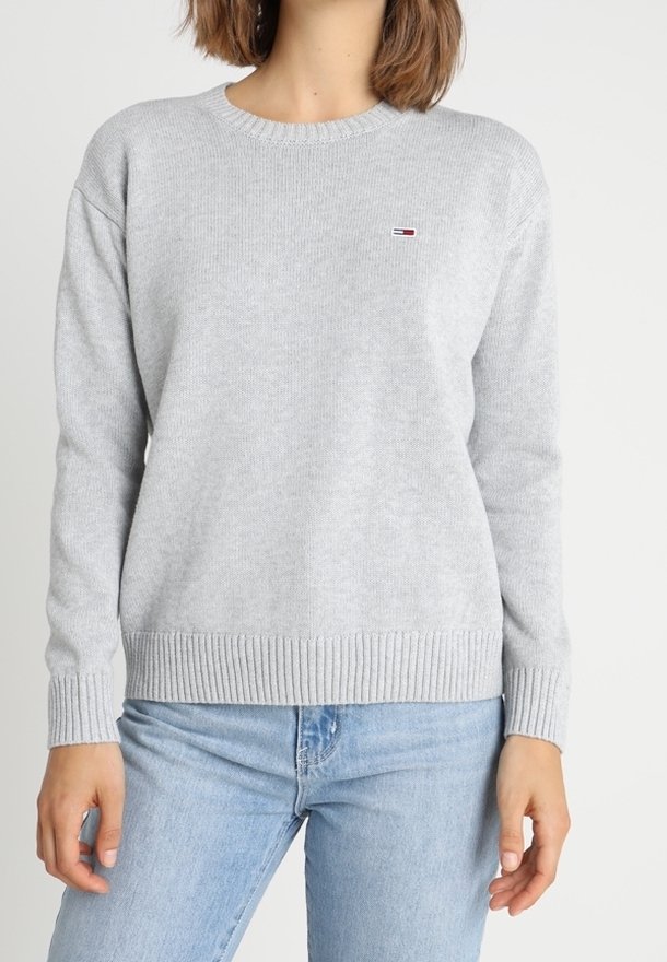 Femme portant un pull en maille gris clair avec un petit logo rouge, blanc et bleu sur la poitrine, associé à un jean bleu clair, debout sur fond blanc.