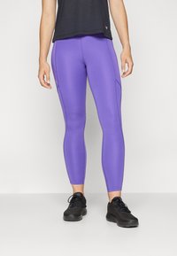 Donna che indossa leggings sportivi viola, scarpe da corsa nere e una maglietta nera, in piedi contro uno sfondo chiaro uniformemente.