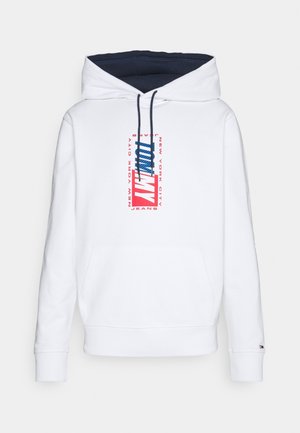 Biała bluza z kapturem, z granatowym wnętrzem kaptura, przednią kieszenią kangurka oraz czerwono-niebieskim nadrukiem „Tommy Jeans New York City” na środku klatki piersiowej.