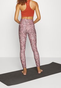 Reggiseno sportivo bordeaux con design testurizzato abbinato a leggings a vita alta con un motivo a macchie rosa e bianche. Scalza su un tappetino da yoga.