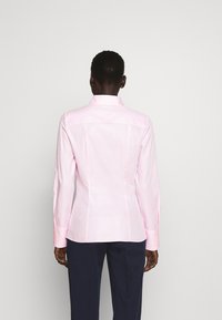Chemise rose clair ajustée avec un col structuré et des manches longues, présentant une texture verticale subtile. Portée avec un pantalon foncé. Vue de dos.