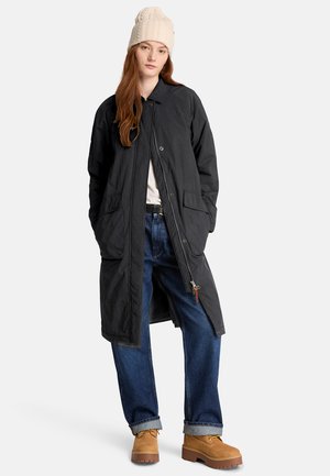 Frau trägt einen langen schwarzen Mantel, blaue Jeans, beige Stiefel und eine cremefarbene Strickmütze, steht vor einem einfarbigen hellen Hintergrund.