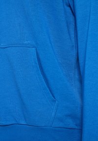 Sweatshirt blu realizzato in morbido tessuto testurizzato, con tasca frontale e orlo a costine. I dettagli delle cuciture sono visibili sulla tasca.