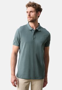 Pikétröja i dämpad teal-färg, kortärm, treknappsknäppning, mjukt bomullsmaterial och subtil logotypdetalj på bröstet.