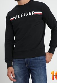 Svart sweatshirt med "HILFIGER"-logo i vitt och röda och blå ränder över axlarna. Mjuk, stickad tyg med ribbade muddar.