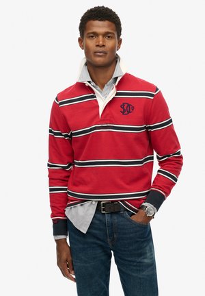 Rotes Langarmpolo mit Navy- und weißen Streifen, Kragen über einem blau-weißen karierten Hemd, Jeans und einer Uhr.