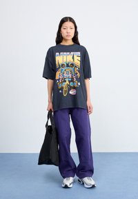 Črna oversized majica s pisanom Nike grafiko, vijolične široke hlače in srebrni športni čevlji. Model drži črno torbo. Enobarvna ozadje.