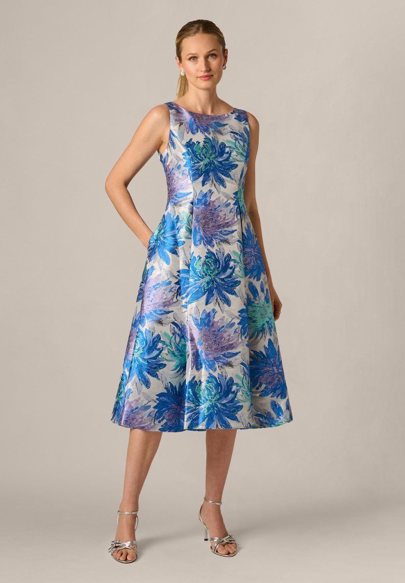 Femme debout portant une robe midi sans manches argentée avec un grand imprimé floral bleu et violet, associée à des sandales argentées à talons hauts.