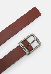 Smeđi kožni remen sa srebrnom metalnom kopčom. Kopča sadrži logotip, a remen ima ravnomjerno raspoređene rupice za prilagodbu.