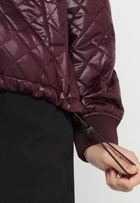 Bordeaux gewatteerde jas met een glanzende textuur, elastische manchetten en een verstelbare koord aan de onderkant. Zwarte achtergrond contrasteert met het kledingstuk.