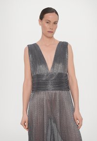 Abito metallico grigio con scollatura a V profonda, texture plissettata e vita aderente. La gonna fluida si drappeggia delicatamente, creando un movimento elegante.