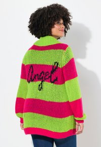 Cardigan en tricot présentant de larges rayures horizontales en rose vif et vert, avec un grand graphique "ange" en noir au dos. Design à col haut.