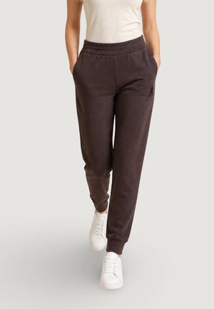 Femme portant un pantalon de jogging noir avec taille et poignets élastiques, des baskets blanches et un haut blanc sans manches, marchant vers l'avant.