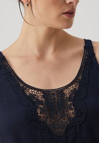Kék tank top, amelyen bonyolult csipkebetétek találhatóak a nyakkivágás körül. Az anyag puha tapintású és sima felületű.