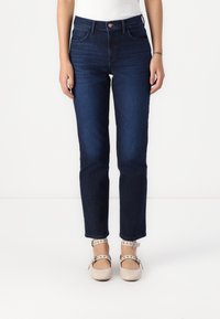 Person wearing dark blue high-waist straight-leg jeans, white top, and beige ankle-strap shoes with buckle and stud details.