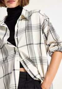 Kvinna i svart polotröja och svarta byxor med en vit och grå rutig skjorta med uppkavlade ärmar.