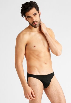 Schwarze Schwimmshorts mit glatter Textur, enganliegendem Design und dezentem Logo-Detail am Bund, Modell posiert mit der Hand am Hals.