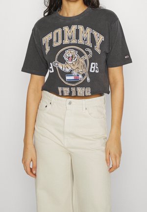 Kobieta nosząca krótki czarny t-shirt Tommy Jeans z grafiką ryczącego tygrysa oraz beżowe spodnie z wysokim stanem.