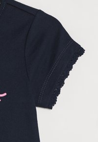Námořnická modrá bavlněná košile s krátkým rukávem, která má krajkový lem. Hladká textura a kontrastní švy. Jednoduchý design bez vzorů.