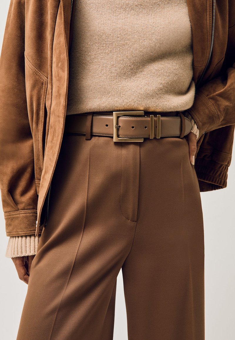 Gros plan d'une personne portant un pantalon marron taille haute avec une ceinture en cuir assortie, un pull beige et une veste en daim marron.