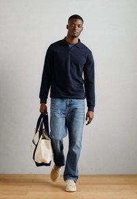 Tengerészkék, hosszú ujjú felső mandarin gallérral, kék farmernadrággal és bézs sportcipővel. Vászontáska sötét részletekkel a kezében.