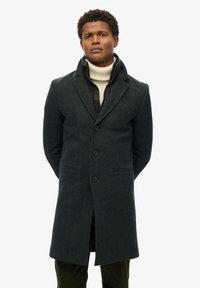 Ciemnoszary wełniany płaszcz o dopasowanym kroju, z klapami i czterema guzikami. Nałożony na czarną pikowaną kurtkę wewnętrzną, z wysokim kołnierzem.