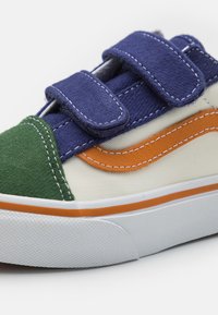 Zapatillas de velcro con acentos en verde, marino y naranja sobre material de lona blanca. Presenta una suela de goma texturizada y costuras en contraste.