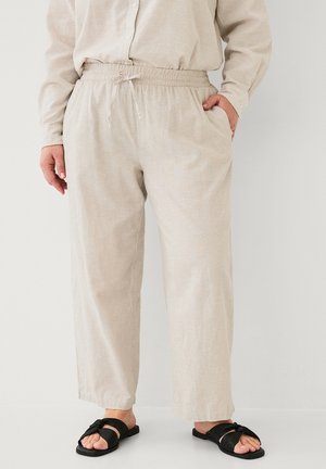 Person iført beige bukser med snor og beige langærmet skjorte, stående med den ene hånd i lommen og sorte slip-on sandaler.