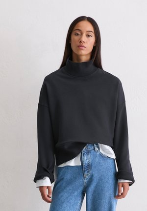 Zwarte cropped sweatshirt met een hoge kraag, geribbelde boorden en een zachte textuur, gedragen over een wit met blauw gestreept shirt en lichtblauwe jeans.