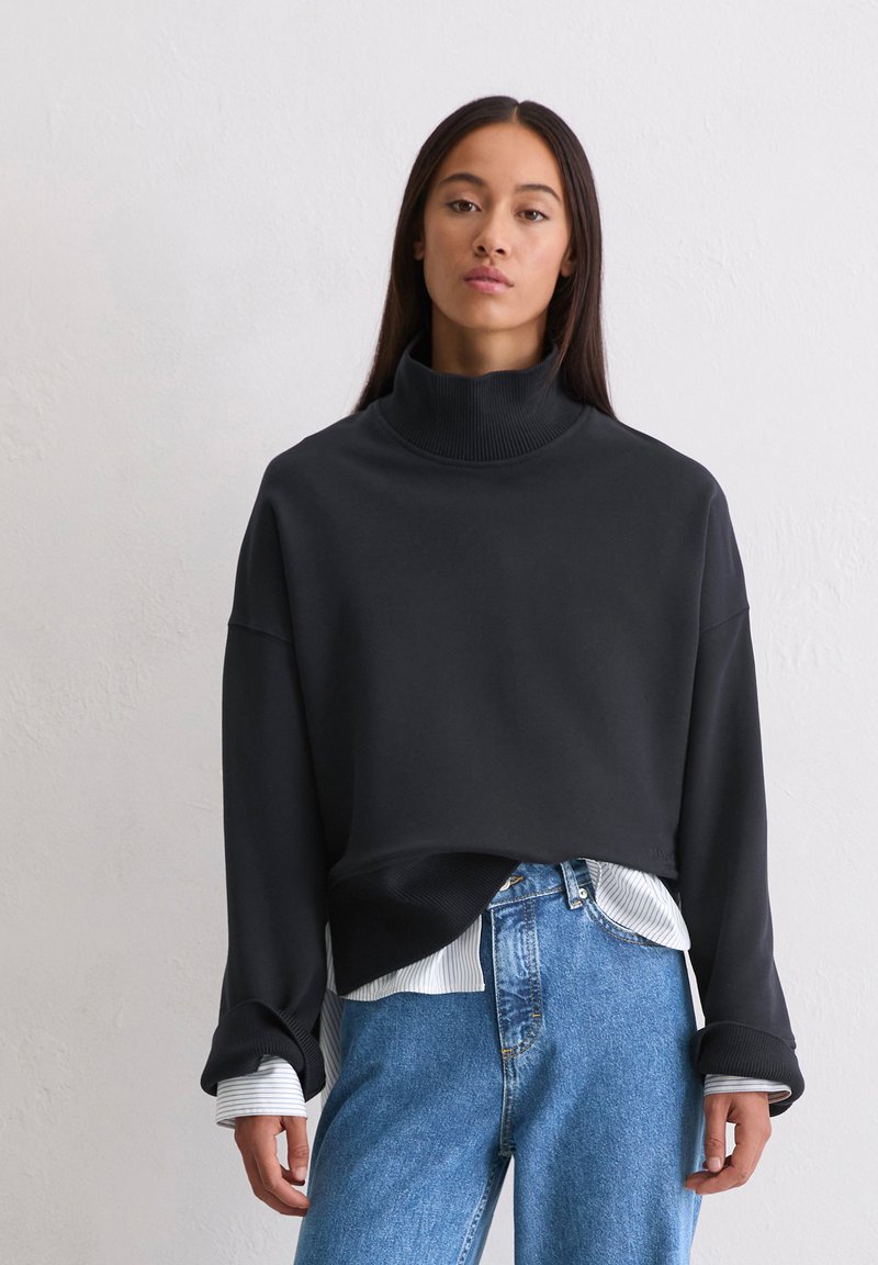 Zwarte cropped sweatshirt met een hoge kraag, geribbelde boorden en een zachte textuur, gedragen over een wit met blauw gestreept shirt en lichtblauwe jeans.