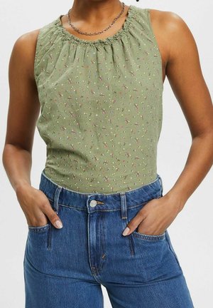 Henkilö pukeutunut vihreään, kukalliseen hihattomaan toppiin, jossa on poimutettu pääntie, ja korkeavyötäröisiin sinisiin farkkuihin, kädet taskuissa.