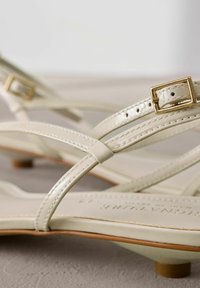 I sandali in vernice beige presentano cinturini incrociati, piccole fibbie dorate e un design a tacco basso con una soletta liscia e opaca.