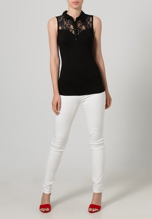 Zwarte mouwloze top met kantdetails op de schouders, knoopsluiting aan de voorkant, gecombineerd met witte skinny jeans en rode sandalen.