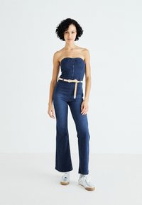Denim kombinezon brez naramnic, temno modre barve in s širokimi hlačnicami. Ima svetlo bež pas okoli pasu. Vključeni so srebrni teniški čevlji.