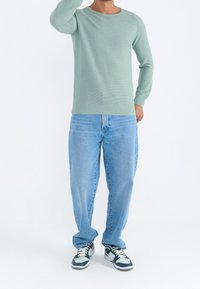 Pull côtelé vert clair à col rond, associé à un jean bleu ample et des chaussures de sport noires avec des accents blancs. Tenue décontractée.