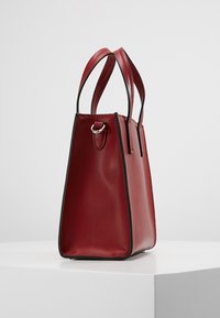 Punainen nahkainen kassi, jossa on kolmiomainen muoto, sileä pinta, mustat korostusvaraosat ja kaksi lyhyttä kantohihnaa.